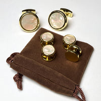 A set of round cufflinks with iridescent mother-of-pearl faces and gold-toned metal hardware, displayed on a brown velvet drawstring pouch against a light gray background.