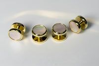 A set of gold-tone ear plugs with round iridescent mother-of-pearl inlays, shown both front and back on a light gray background.
