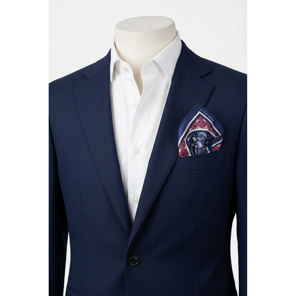 A navy blue suit jacket displayed on a cream mannequin torso, paired with a crisp white dress shirt and a patterned pocket square in navy, burgundy, and gold.