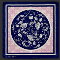 A square scarf laid flat on a gray textured fabric background. The scarf features a navy blue border, a central navy blue circle with white floral and vine motifs, a pink inner field with white floral patterning, and the brand name 'A.