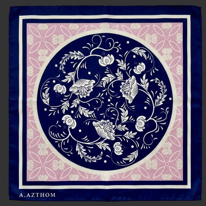 A square scarf laid flat on a gray textured fabric background. The scarf features a navy blue border, a central navy blue circle with white floral and vine motifs, a pink inner field with white floral patterning, and the brand name 'A.