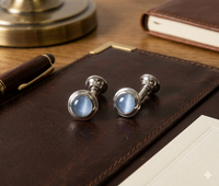 A pair of silver-toned cufflinks with round light blue cabochon gemstones, displayed on a dark brown leather notebook cover on a wooden desk.