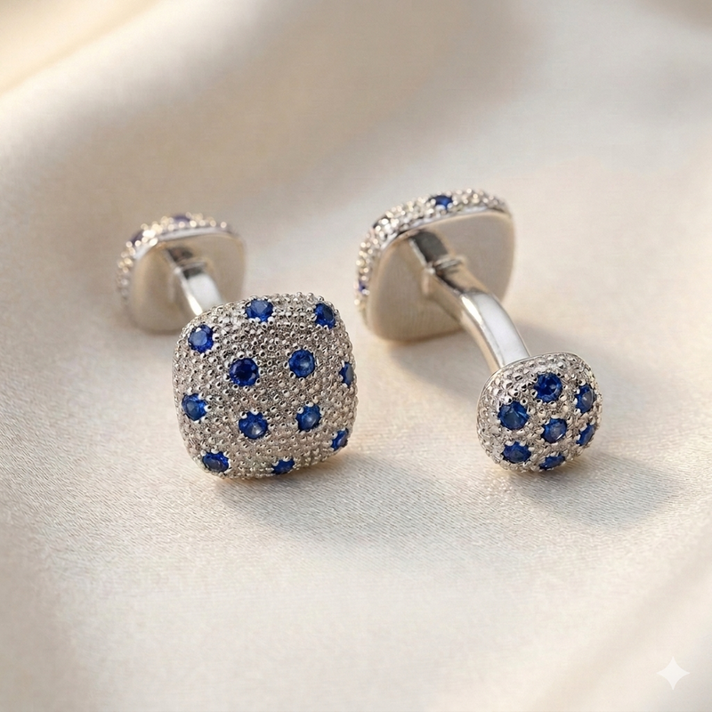 A pair of silver-toned cufflinks with square faces covered in small blue gemstones, photographed on a soft cream fabric background.